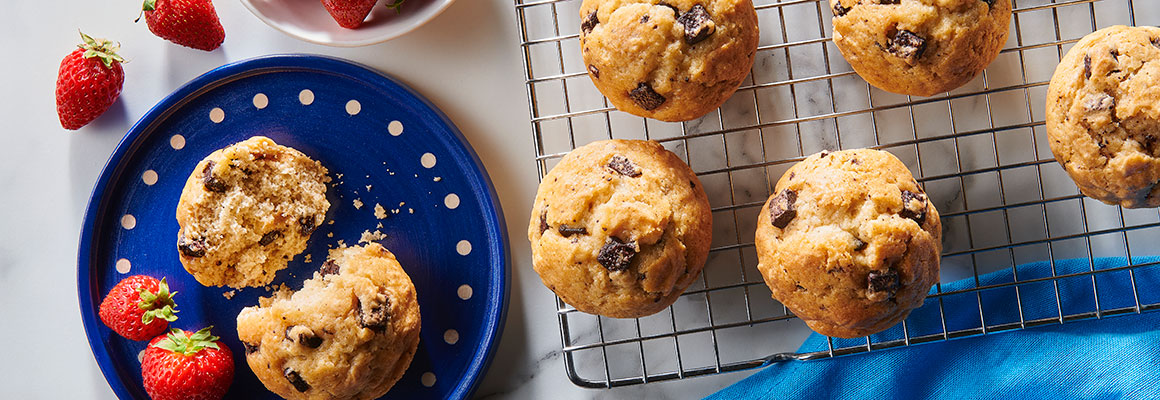 Top view of Pillsbury muffins on a cooling rack and a blue barrel head dotted plate. The plate also contains a halved muffin and fresh strawberries. A bowl of whole strawberries and a blue napkin are visible on the white marble countertop.