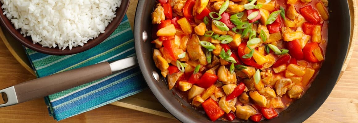 A skillet of 5-Ingredient Sweet-and-Sour Chicken stir-fry with bell peppers and green onions. Served with a bowl of white rice and a small dish of peanuts on a wooden tray.