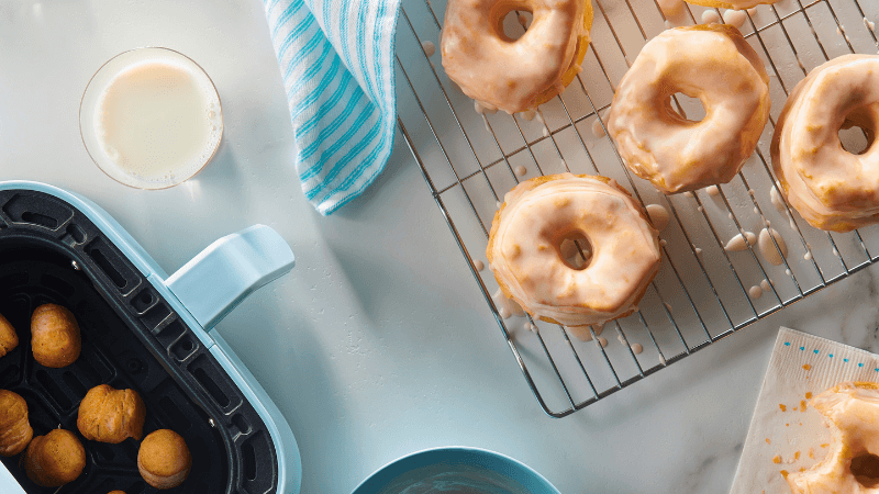 Freshly glazed doughnuts cooling on a wire rack with doughnut holes frying in an air fryer nearby.